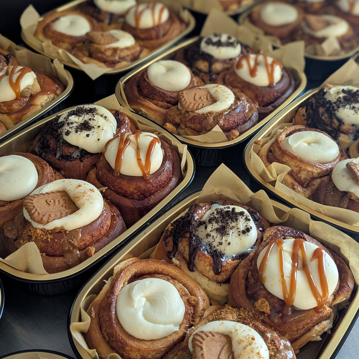 Multiple trays of assorted cinnamon rolls filled with white frosting, drizzled with caramel sauce, and topped with biscuit pieces, photographed on a dark surface.