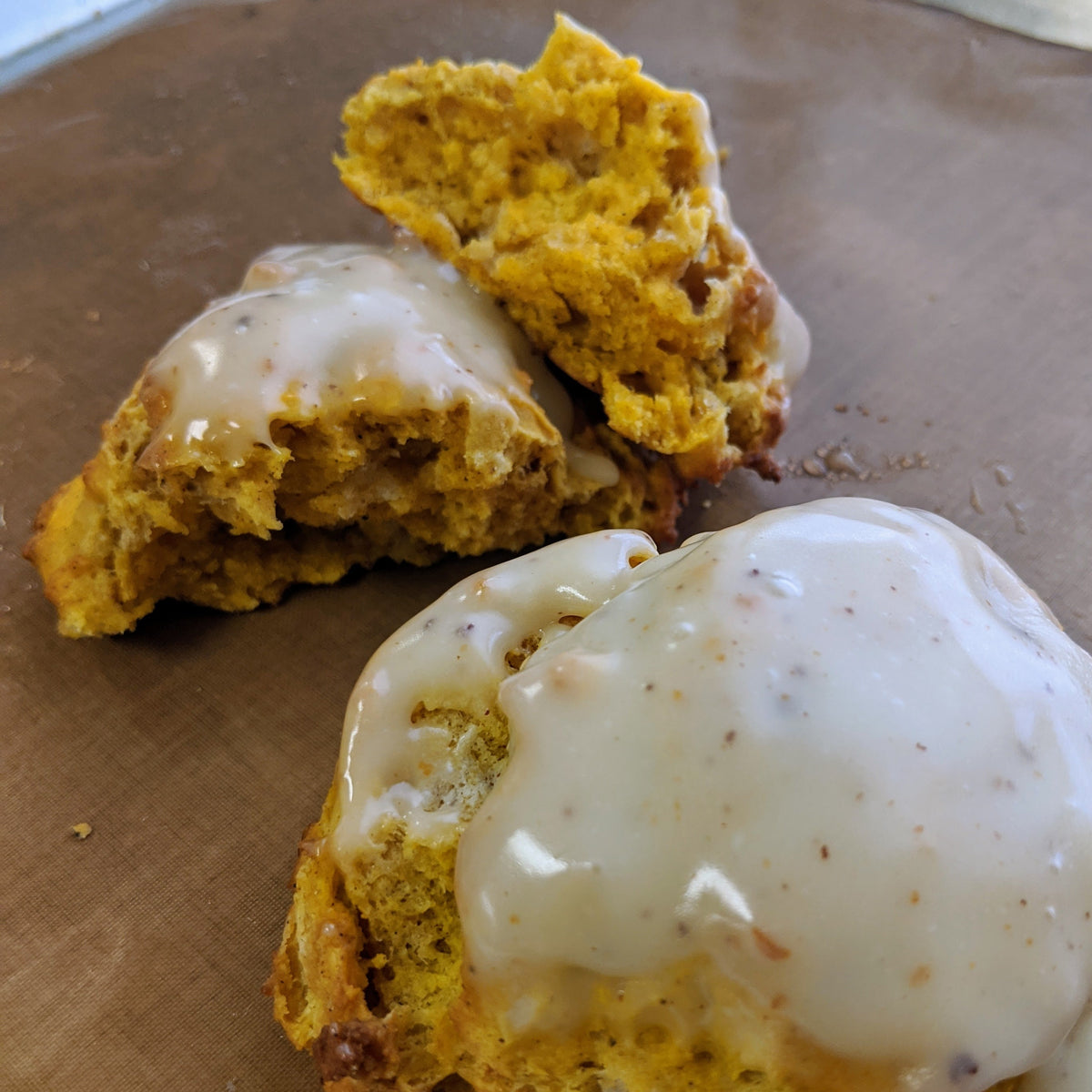 Two golden-brown pumpkin scones with white glaze, one cut open to show the crumb, on a brown baking sheet.