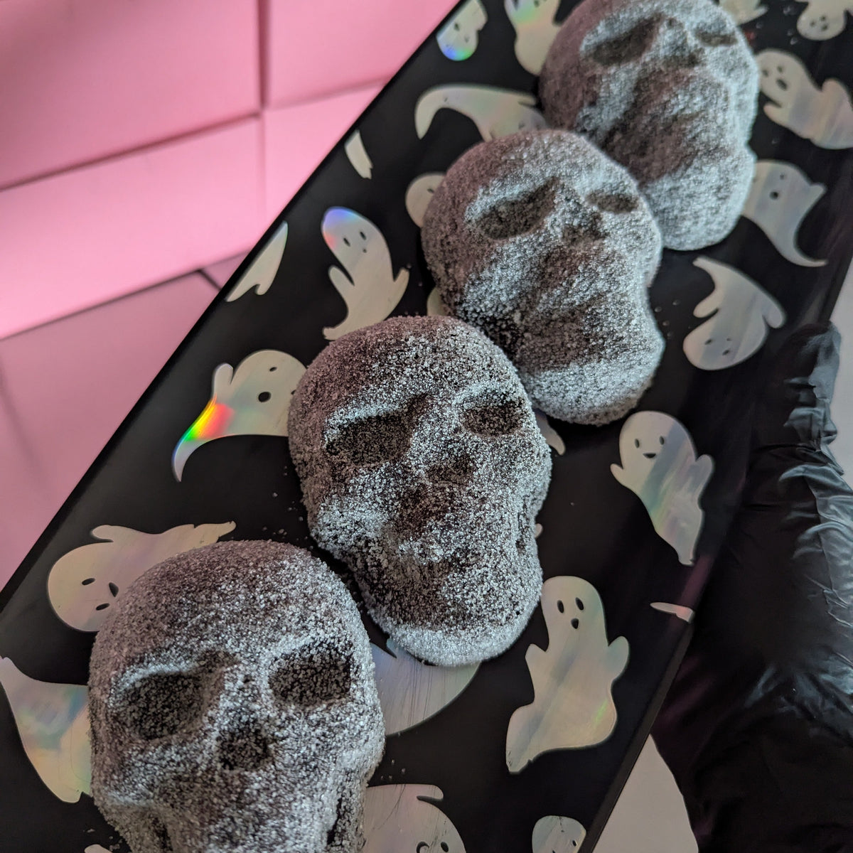 Four sugar-dusted skull-shaped treats arranged on a black tray with white ghost illustrations, held by a hand wearing a black glove against a pink background.