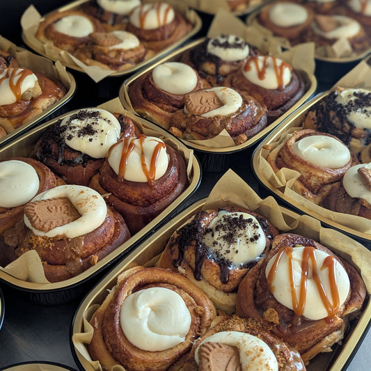 Multiple trays of assorted cinnamon rolls filled with white frosting, drizzled with caramel sauce, and topped with biscuit pieces, photographed on a dark surface.