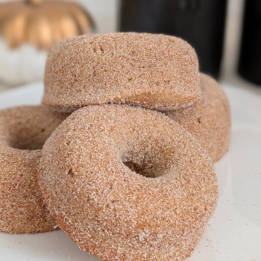 A pile of sugar-coated donuts on a white surface, with a black candle and a small gold pumpkin blurred in the background.